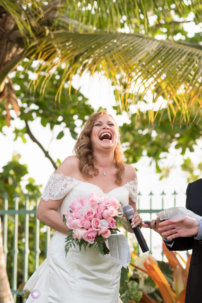 Bride wearing a white dress holding a bouquet of pink roses laughs and smiles while holding the microphone during her ceremony.