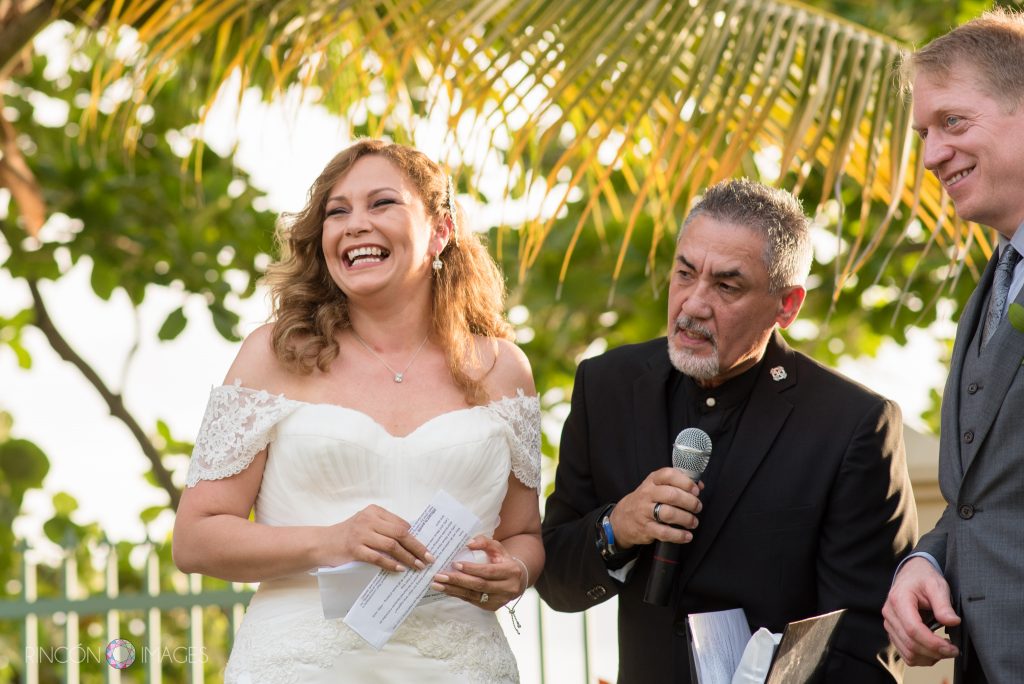 The bride, groom and minister all smiling as the bride begins to unfold her vows. They are stading in front of plam trees with yellow and green frauns.
