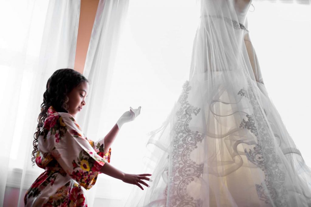 Wedding photograph of a flower girl wearing a floral robe touching a wedding dress hanging in a window.
