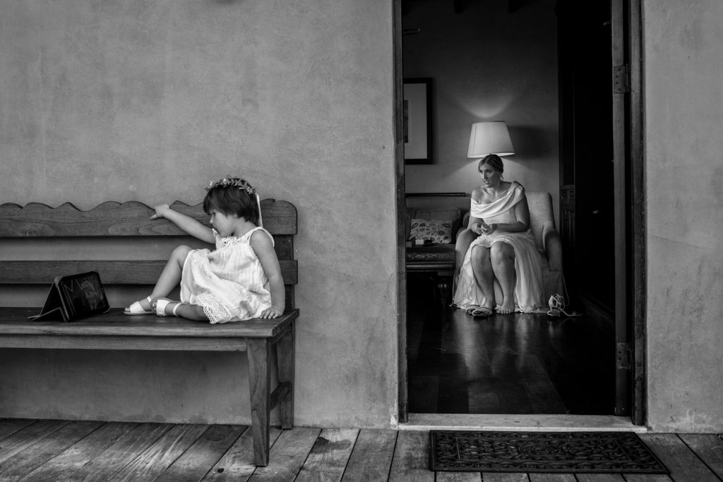 Flower girl sitting on a bench watching an ipad at wedding with a bridesmaid in the background.