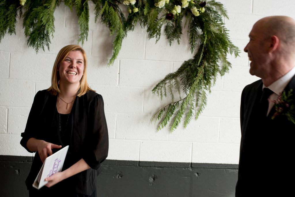 Officiant and groom laughing in front of pine foliage and white flower wedding decorations.