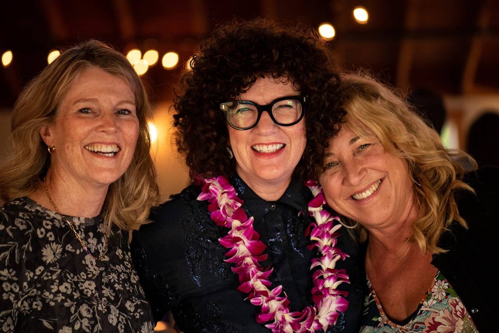 Picture of three wedding guests smiling while one is wearing a lei made of orchid flowers at the reception at The Loft at The Red Building in Astoria, Oregon.